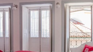 Un dormitorio con colcha roja, suelo de madera y una silla blanca con cojín rojo.
