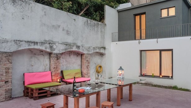 Un patio al aire libre con una mesa de vidrio, taburetes de madera y un sofá colorido.
