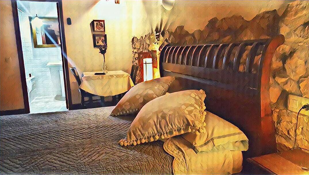 A bedroom with a stone wall, a bed with a headboard, two pillows, a small table, and a chair.