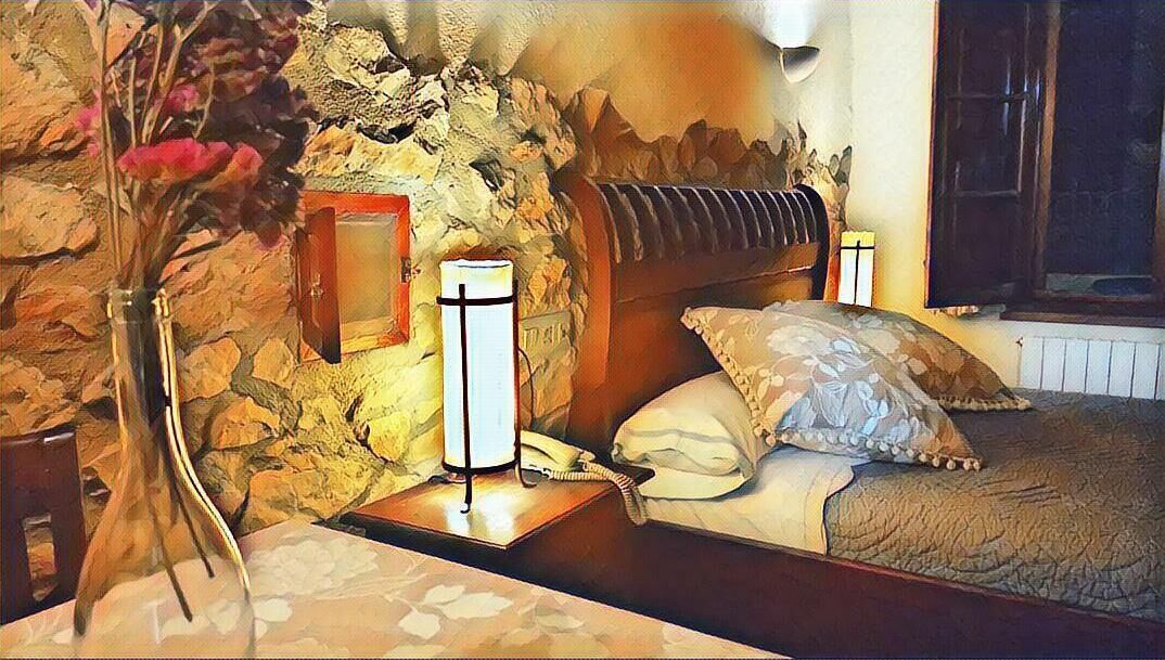 Un dormitorio con pared de piedra, una cama con colcha estampada, una mesita de noche con lámpara y un florero con flores.