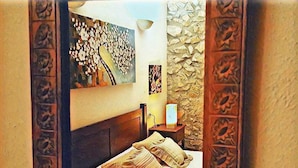 Une chambre à coucher avec une tête de lit en bois, un lit avec une literie beige, une table de chevet avec une lampe, et un mur recouvert d’un revêtement en pierre texturée et orné d’un tableau floral.