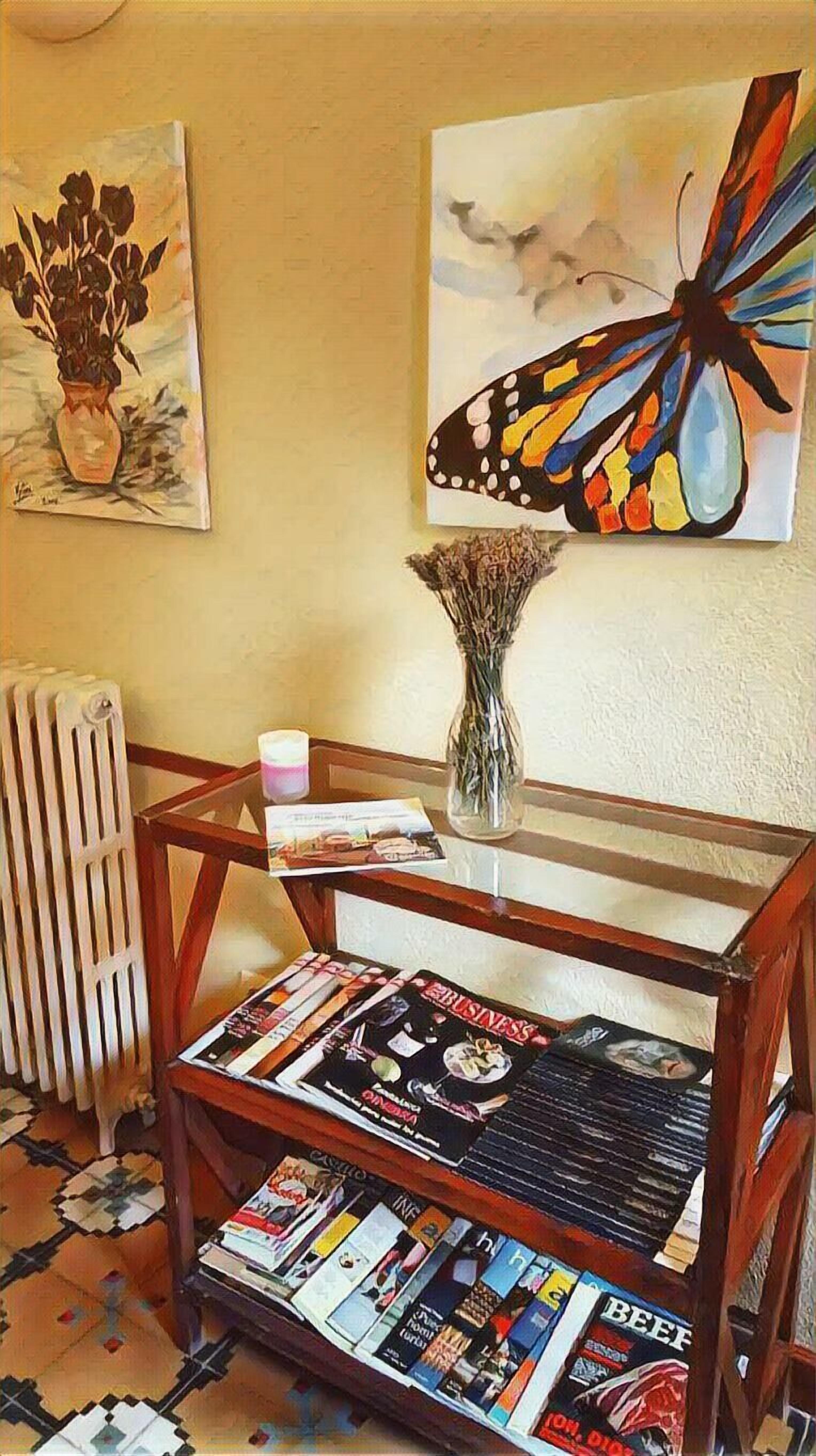 A room with a glass-top table displaying magazines, a vase with dried flowers, and two paintings on the wall.