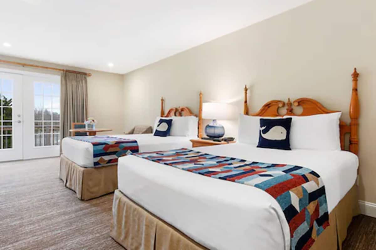 A hotel room with two beds, a large window with curtains, a wooden headboard, and a small table with a lamp.