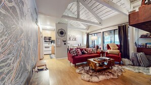 Een ruime woonkamer met een rode bank, een houten salontafel en een bont tapijt. In de achtergrond is een keuken met moderne apparatuur zichtbaar.