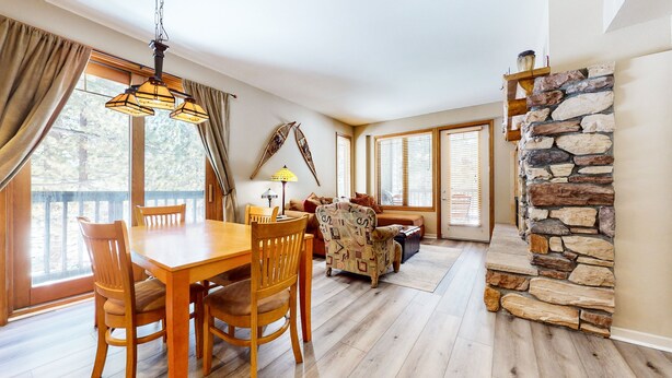 A spacious living area with a wooden dining table and chairs, a patterned armchair, a stone fireplace, and large windows with blinds. - Mammoth Green #111 2 Bedroom Condo (Mammoth Lakes)