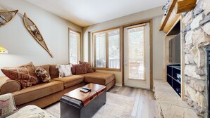 A living room with a brown sectional sofa, a coffee table, and a stone fireplace. - Mammoth Green #111 2 Bedroom Condo (Mammoth Lakes)