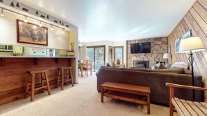 A cozy living room with a wooden bench, a sofa, and a fireplace. There is a kitchen area with a counter and stools. - Snowcreek #39 1 Bedroom Townhouse (Mammoth Lakes)
