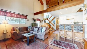 A cozy living room with a brown leather sofa, a coffee table, and patterned rugs. A staircase leads to an upper level with wooden beams. - Snowcreek #638 3 Bedroom Condo (Mammoth Lakes)