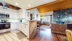 A kitchen with white cabinets, a stove, and a microwave. There is a wooden island with a sink and a chair. The living room has a brown couch and a stone wall with a ship model. - Snowcreek #638 3 Bedroom Condo (Mammoth Lakes)