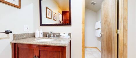 Un baño con lavamanos de encimera de granito, armarios de madera, un espejo grande y una puerta de madera.