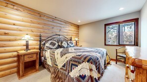 Ein gemütliches Schlafzimmer mit einer Holzwand, einem großen Bett mit einer Stegdecke, zwei Nachttischlampen und einem Fenster mit Blick auf Bäume und Gebäude.