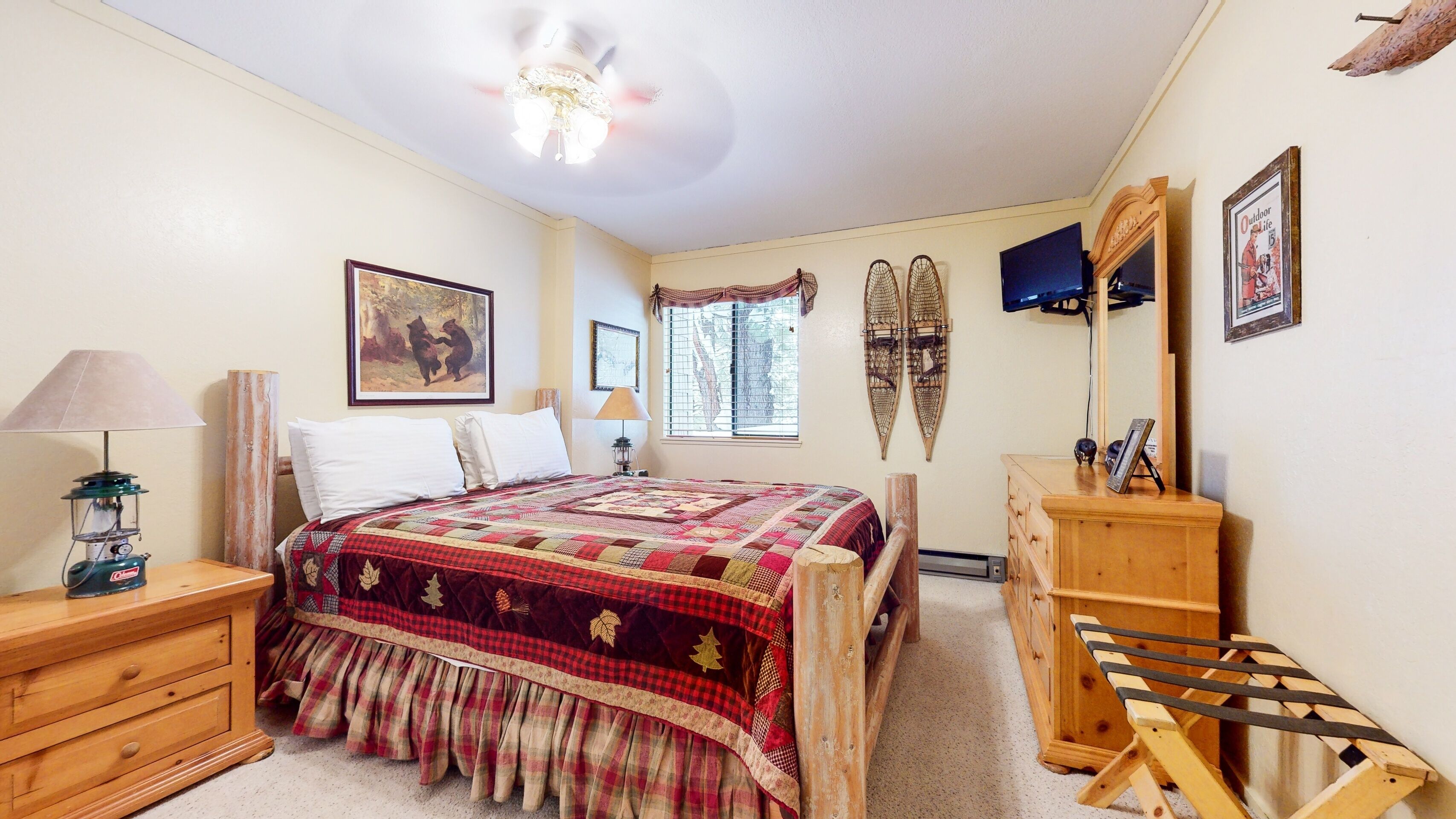 A bedroom with a large bed, a wooden dresser, a desk, a chair, a lamp, a window with a view of trees, and a ceiling fan.
