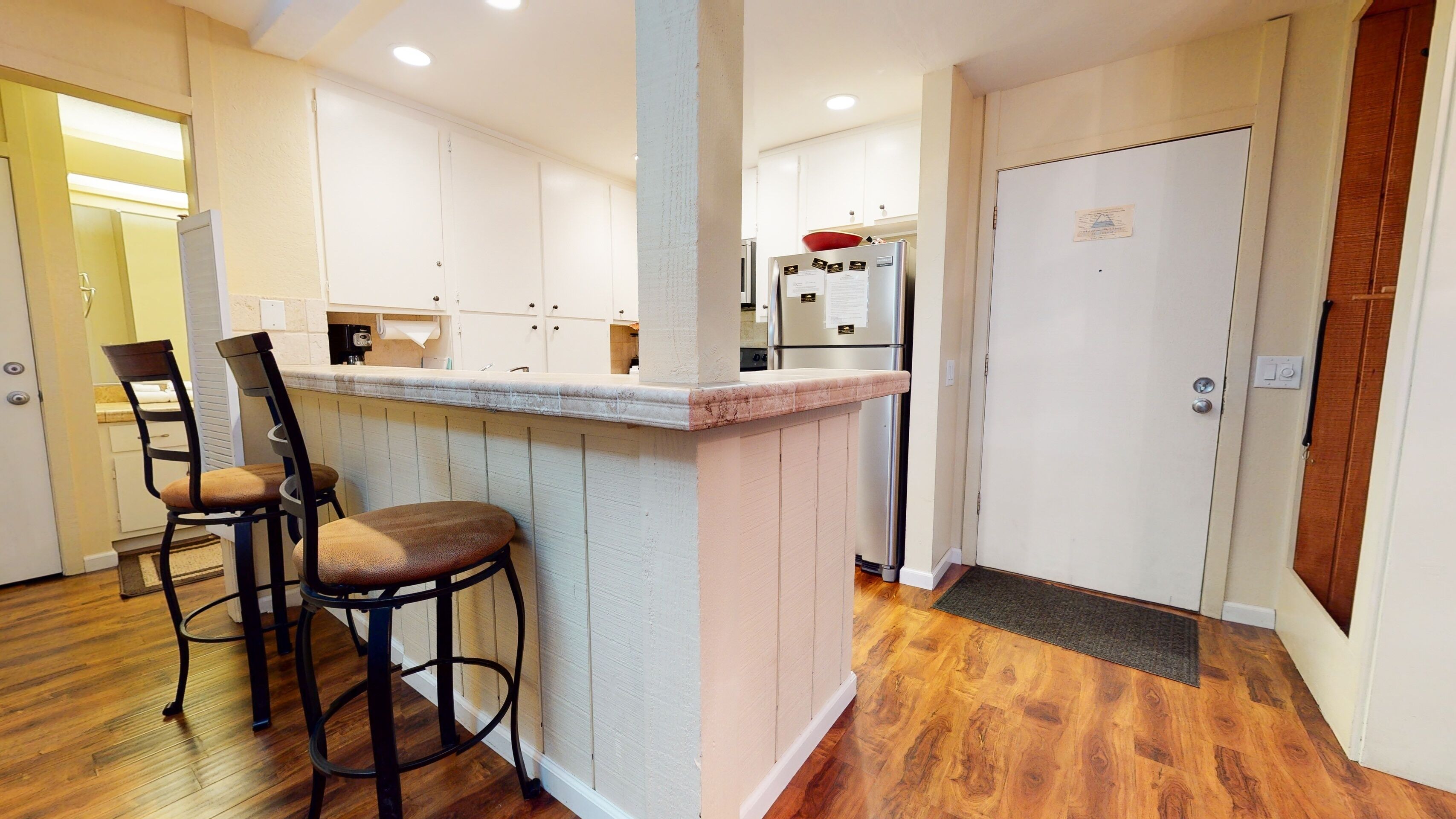 A kitchen with a wooden bar counter, two bar stools, a refrigerator, and a door with a sign.