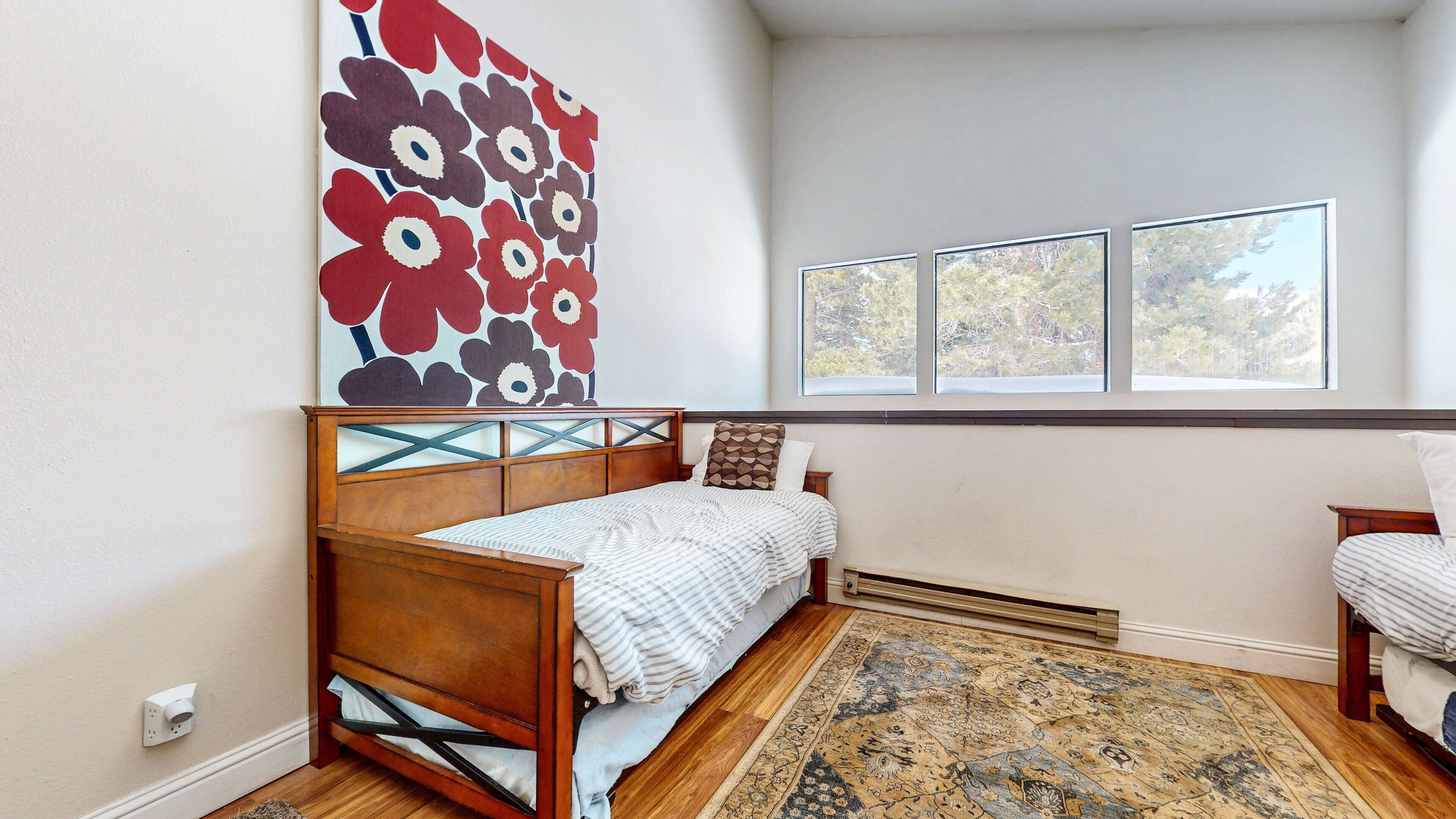 Ein Schlafzimmer mit einem hölzernen Bett, einem gemusterten Teppich, einem großen floralen Kunstwerk an der Wand und einem Fenster mit Blick auf Bäume.
