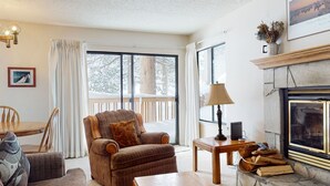 Un salon chaleureux avec une cheminée, un fauteuil confortable, une table à manger avec des chaises et de grandes fenêtres donnant sur des arbres couverts de neige.