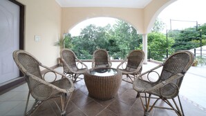 A covered outdoor seating area with wicker furniture and a central table, overlooking a lush garden. - Kangs Nirvana Resorts and Spa (Garhshankar)