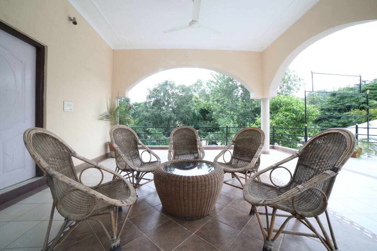 Un espace extérieur aménagé, abrité et doté de mobilier en osier et d’une table centrale, offrant une vue sur un jardin luxuriant.