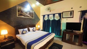 Habitación de hotel con cama, televisión, escritorio, lámpara, un cuadro y ventana con cortinas.