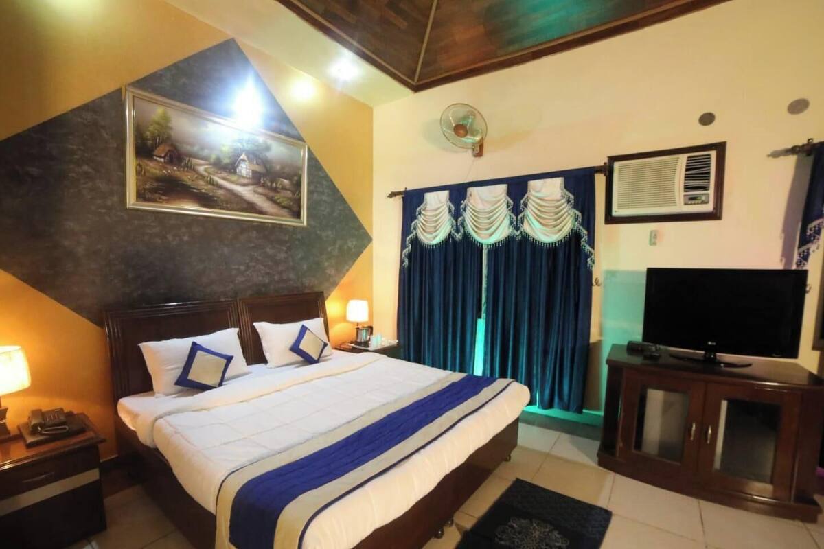 Une chambre d’hôtel avec un lit, une télévision, un bureau, une lampe, un tableau et une fenêtre avec des rideaux.