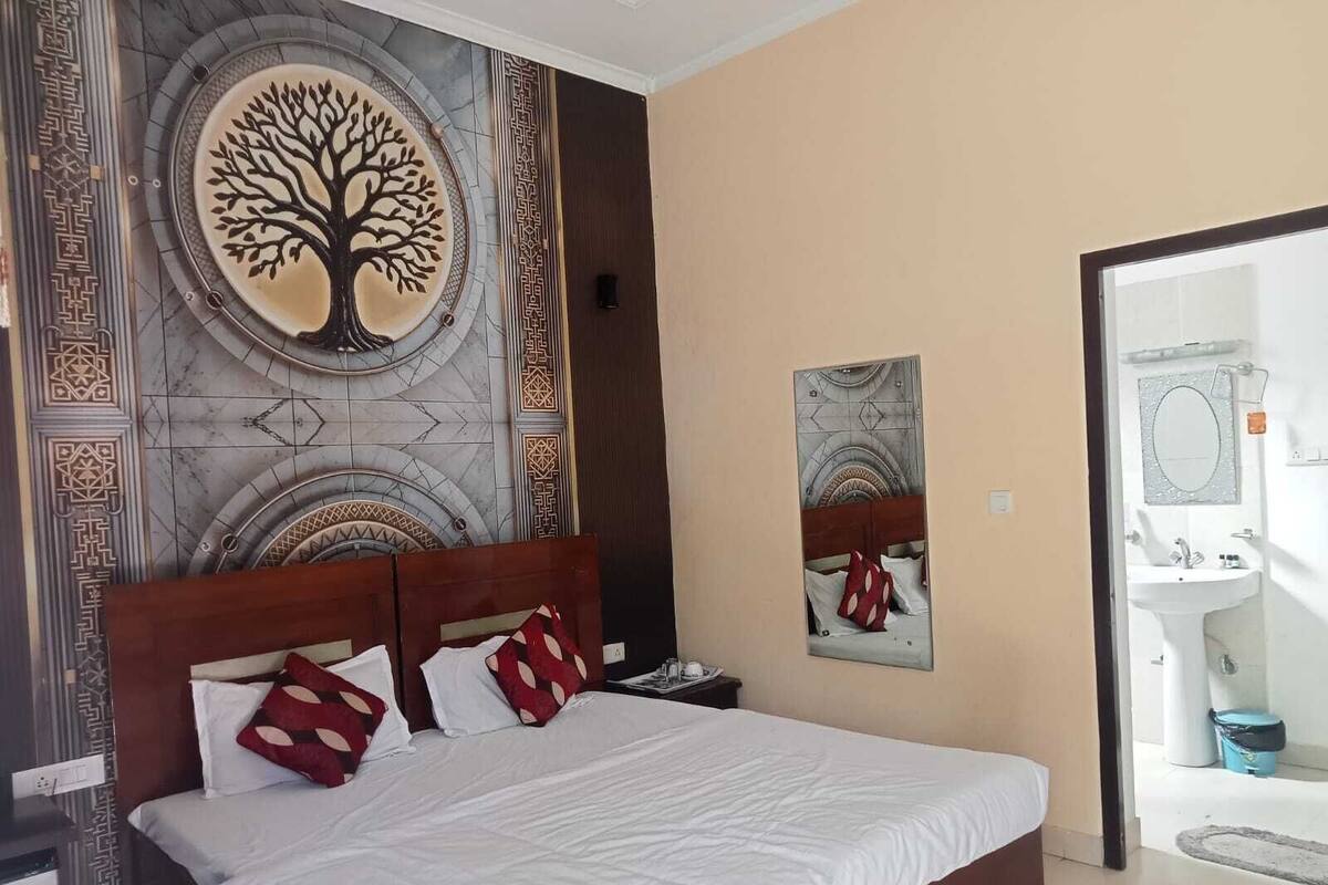 Une chambre d’hôtel avec un lit, une table de chevet, un miroir fixé au mur et une salle de bain visible à travers une porte ouverte.