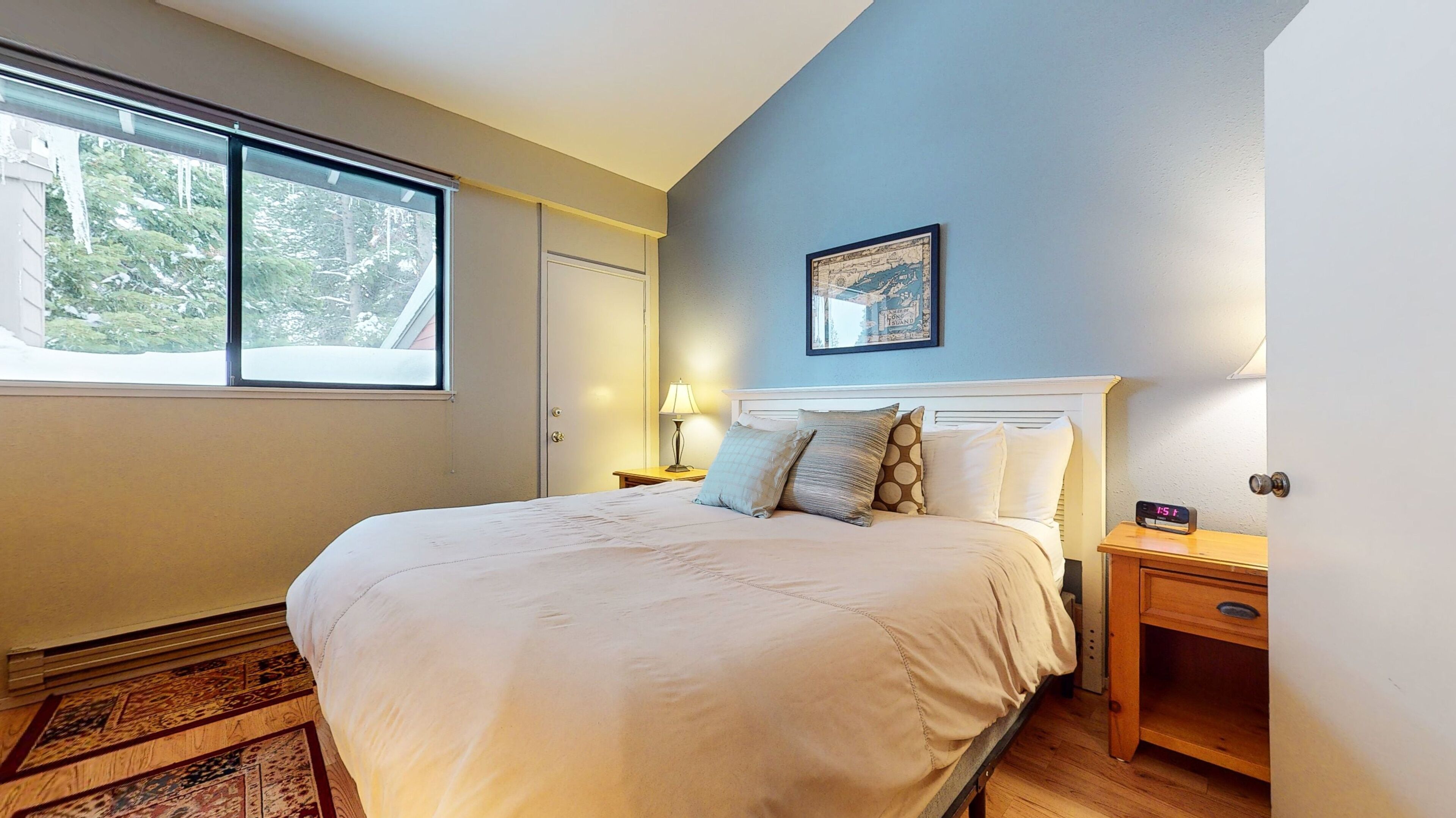 Un dormitorio con una cama grande, una mesita de noche con lámpara, un reloj de pared y una ventana con vistas a árboles.