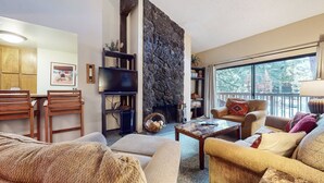 A cozy living room with a stone fireplace, comfortable seating, and a large window offering a view of the outdoors.