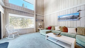 A cozy living room with a green sofa, a white coffee table, and a rocking chair. There is a large window providing a view of trees and a mountain. - Summit #98 3 Bedroom Condo (Mammoth Lakes)