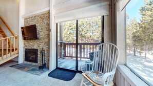 A cozy room with a stone fireplace, a wooden staircase, and a large sliding glass door leading to a balcony with a view of trees. - Summit #98 3 Bedroom Condo (Mammoth Lakes)