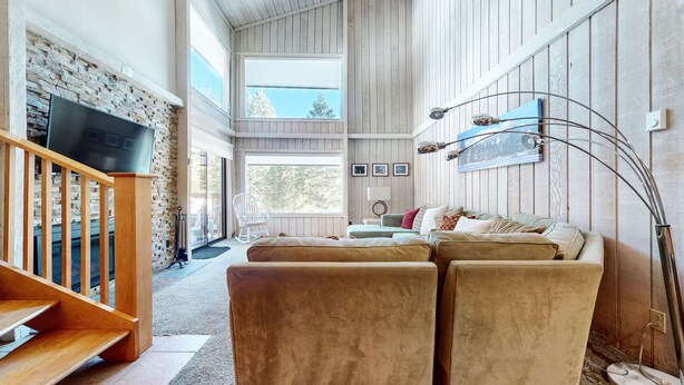 A spacious living room with a stone wall, a wooden staircase, and a large window offering a view of trees. - Summit #98 3 Bedroom Condo (Mammoth Lakes)