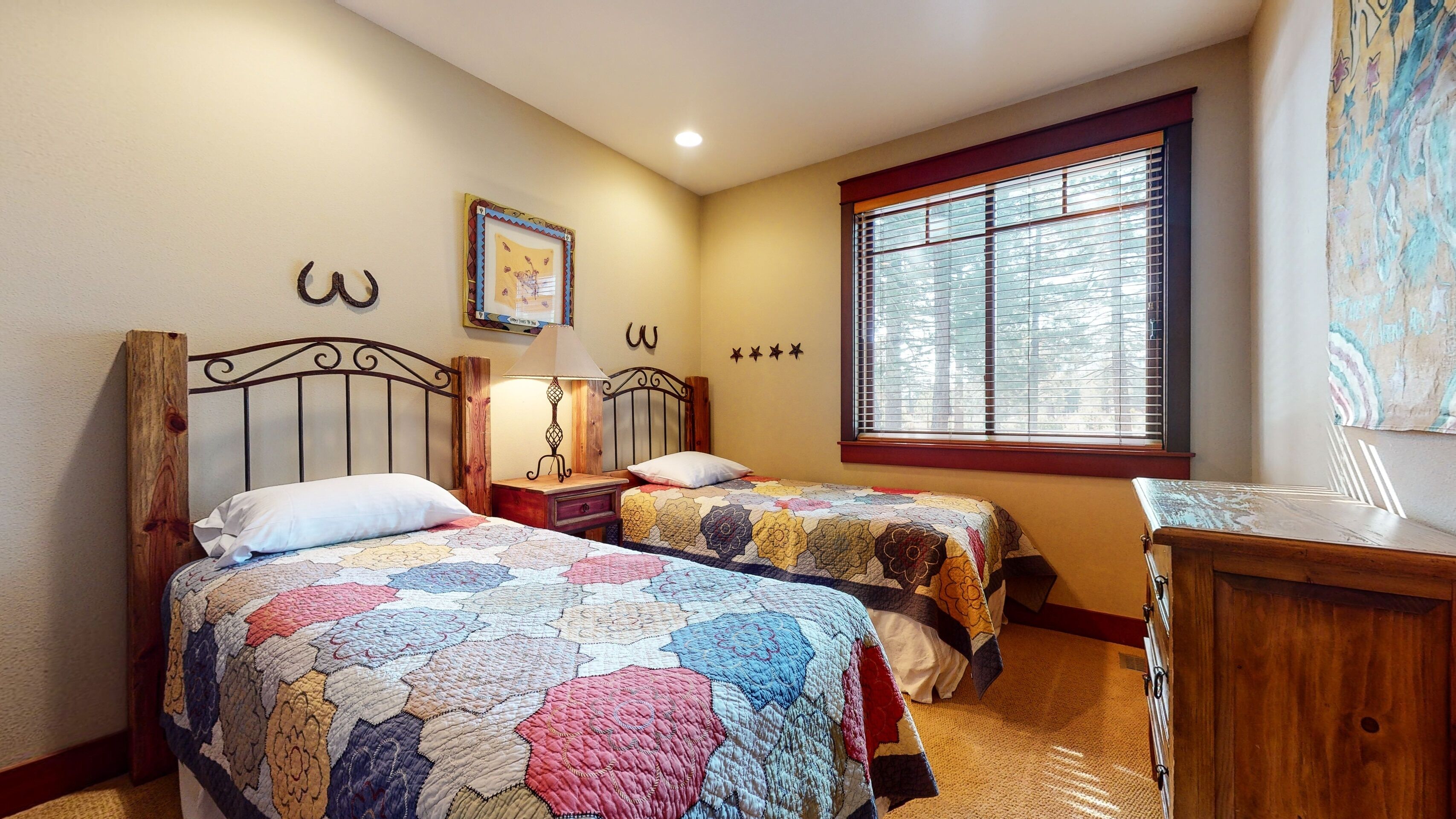 A bedroom with two beds, a wooden headboard, a nightstand with a lamp, a dresser, and a window with blinds.