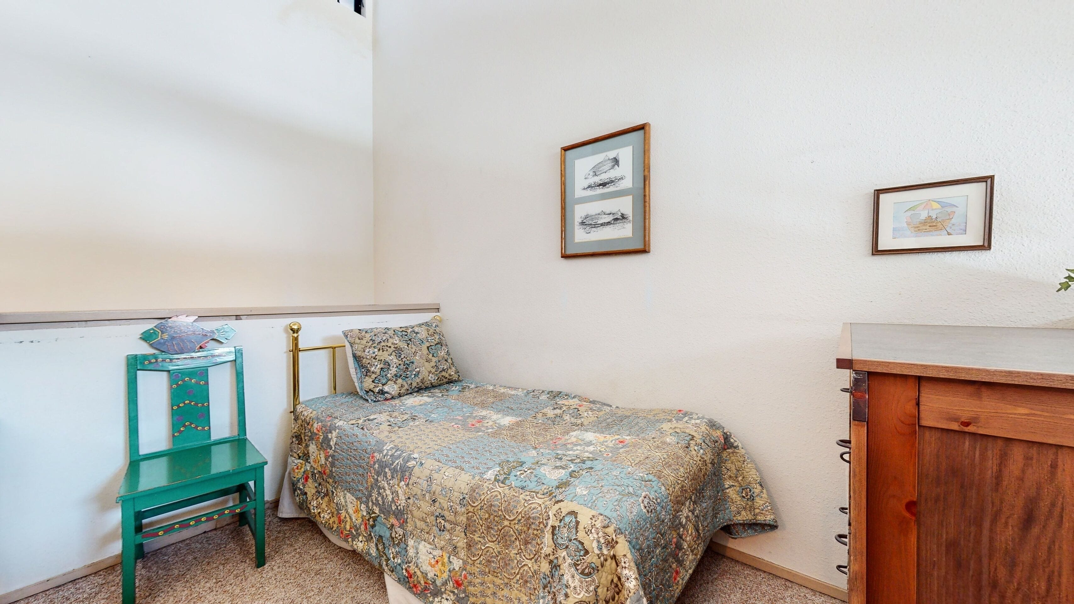 A small, neatly arranged bedroom with a bed, a green chair, a wooden cabinet, and two framed pictures on the wall.