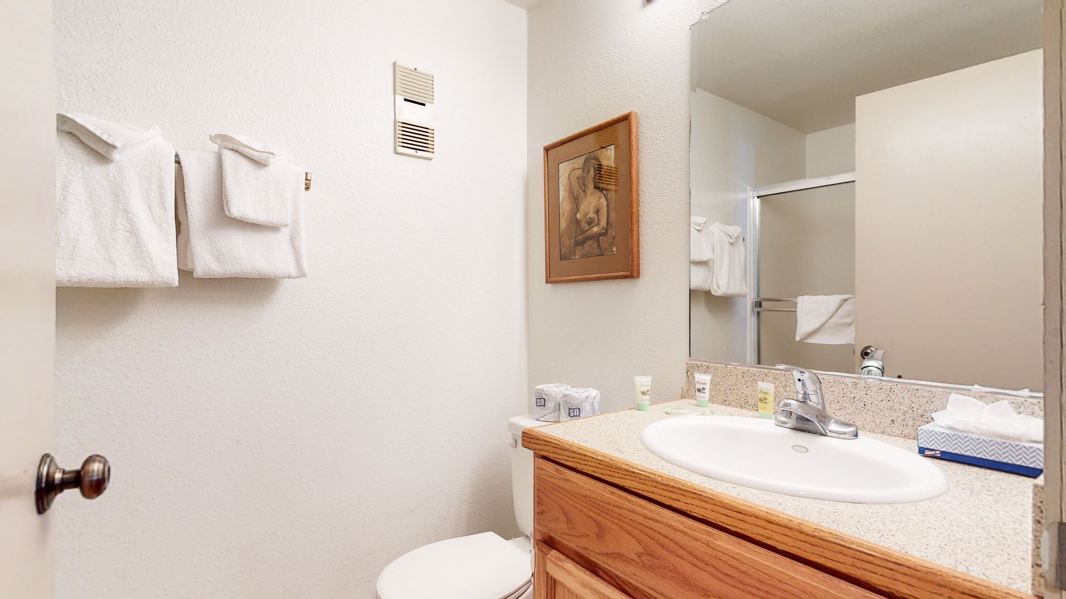 Un baño con un lavabo blanco, tocador de madera y un espejo grande. Hay toallas colgadas en la pared y un cuadro enmarcado sobre el espejo.