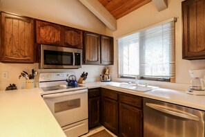 Una cocina con armarios de madera, electrodomésticos de acero inoxidable, encimera blanca y ventana con persianas.