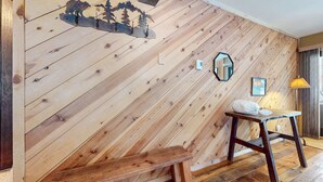 A room with a wooden slat wall, a wooden bench, a small table with a white cushion, a mirror, and a lamp.