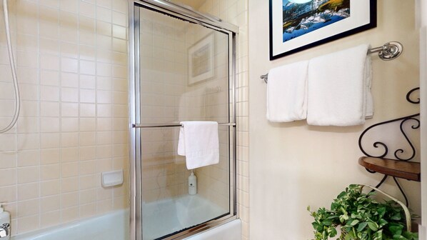 A bathroom with a glass shower enclosure, a white bathtub, a framed picture on the wall, and a potted plant.