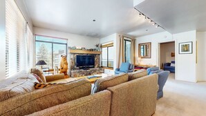 Ein geräumiges Wohnzimmer mit Kamin aus Stein, Fernseher und großem Fenster mit Jalousien.