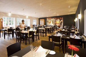 A restaurant with multiple tables and chairs, a woman standing near the entrance, and a large painting on the wall. - Heidehotel Gut Landliebe (Südheide)