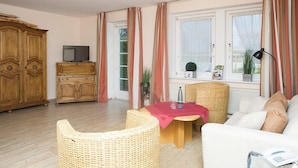 Un salón con un sofá, una silla de mimbre, una mesita con un mantel rojo, un televisor sobre un mueble de madera y una ventana con cortinas.