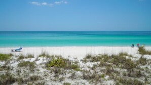 A beach with white sand, clear turquoise water, and sparse vegetation. - Waters Edge 110n 2 Bedroom Condo by RedAwning (Holmes Beach)