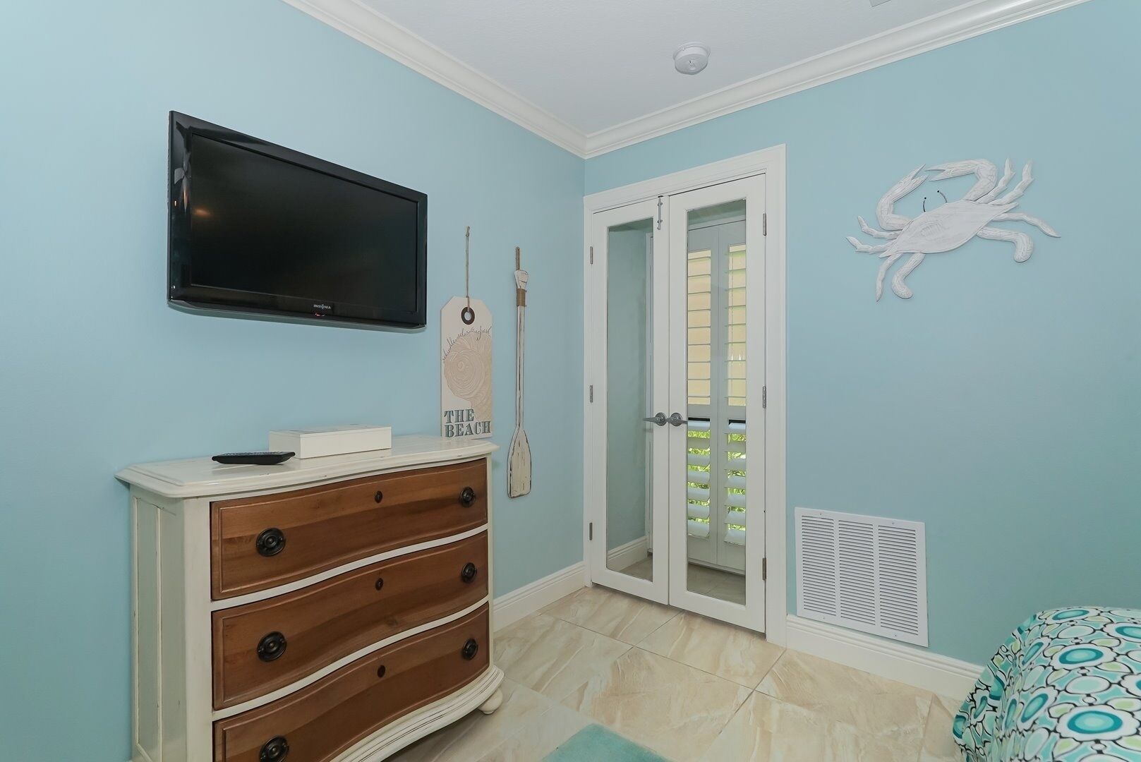 A room with a flat-screen TV mounted on the wall, a wooden dresser with drawers, a door with glass panels, and a decorative crab wall art piece.