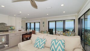 A spacious living area with a beige sofa, two patterned cushions, a dining table with chairs, and a view of the ocean through large sliding glass doors. - Waters Edge 110n 2 Bedroom Condo by RedAwning (Holmes Beach)
