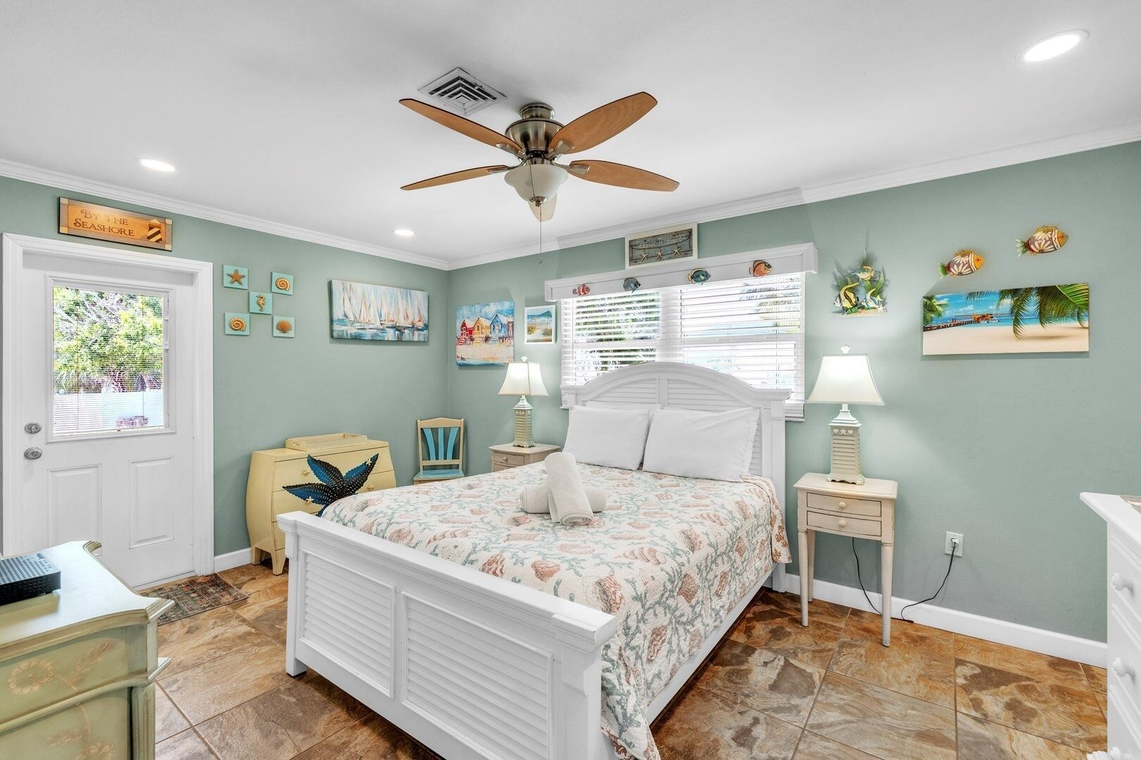 Une chambre à coucher avec un lit, un ventilateur de plafond, une fenêtre et des œuvres d’art accrochées aux murs.