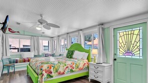 A room with a bed, a chair, a desk, a ceiling fan, and a door with a stained glass window. - Marmalade Mermaid 3 Bedroom Home by RedAwning (Bradenton Beach)