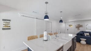 A modern kitchen with a large island, two pendant lights, a sink, and a dining area with wicker chairs.