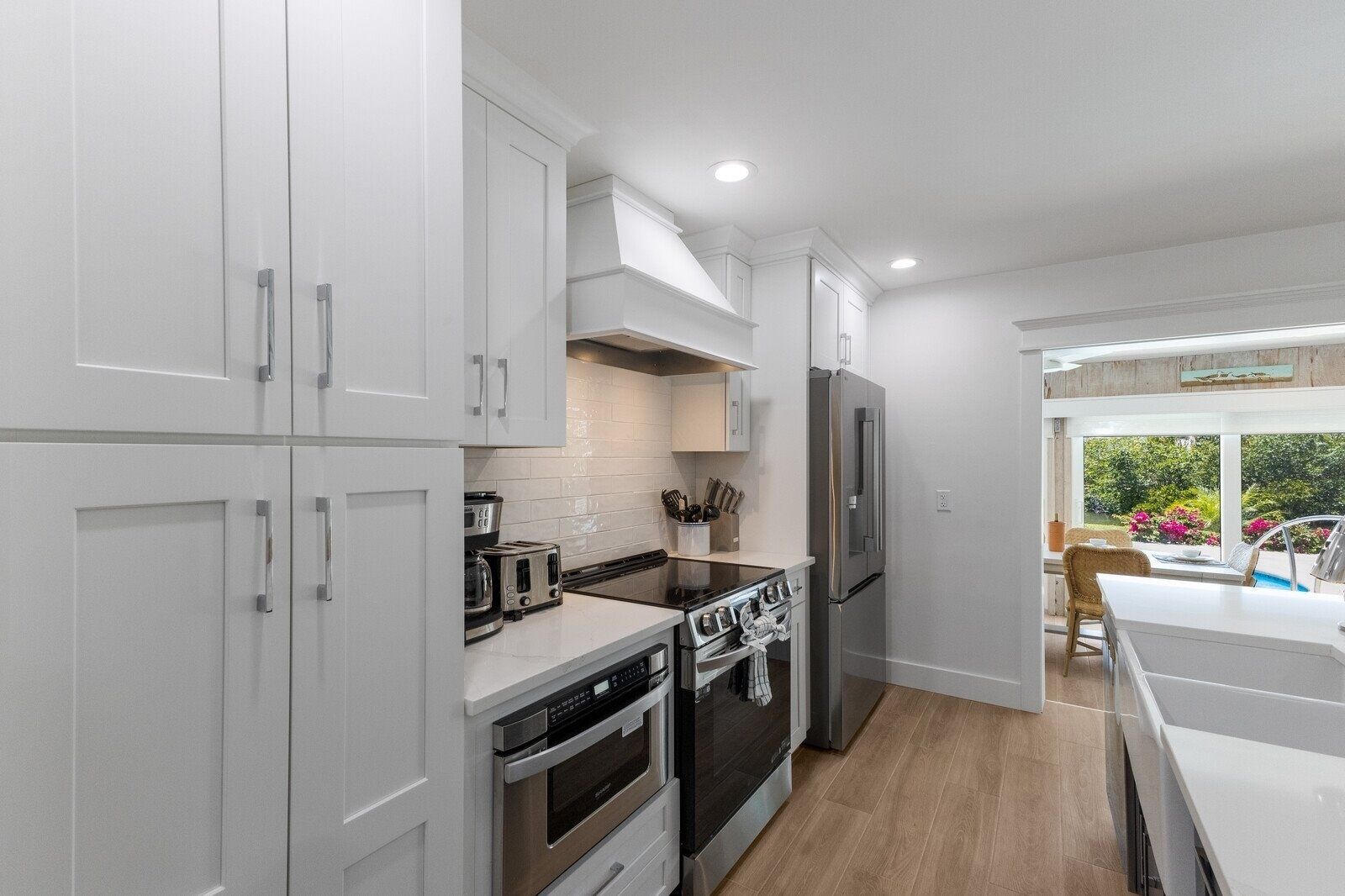 A modern kitchen with white cabinetry, stainless steel appliances, and a view of the garden through a large window.