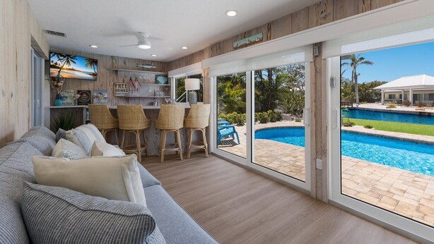 A spacious living area with a large window overlooking a swimming pool, a bar area with wicker chairs, and a cozy seating area with a sofa.