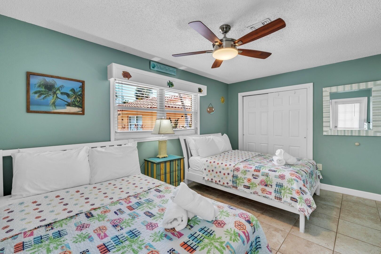 Une chambre avec deux lits, un ventilateur de plafond, une fenêtre avec des stores, une petite table avec une lampe et un miroir fixé au mur.