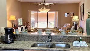 A kitchen with granite countertops, a coffee maker, and a dishwasher. There is a dining area with wicker chairs and a table, and a window with blinds. - Playa Encantada 214 2 Bedroom Condo by RedAwning (Holmes Beach)