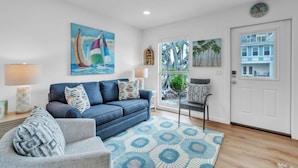 A living room with a blue sofa, patterned cushions, a blue rug, a painting of sailboats, a window with a view of palm trees, and a door with a clock. - Minnow 1 1 Bedroom Cottage by RedAwning (Anna Maria)