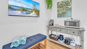 A room with a television displaying a beach scene, a coffee maker, a microwave, a bench with towels, and a shelf with kitchen items. - Minnow 1 1 Bedroom Cottage by RedAwning (Anna Maria)